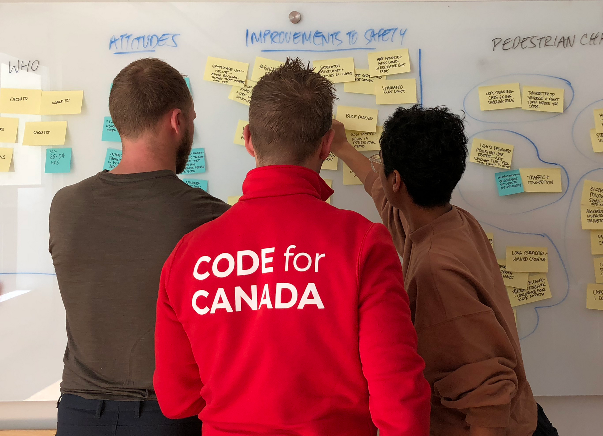 Technologist in Code for Canada branded jacket looks at whiteboard with 2 colleagues