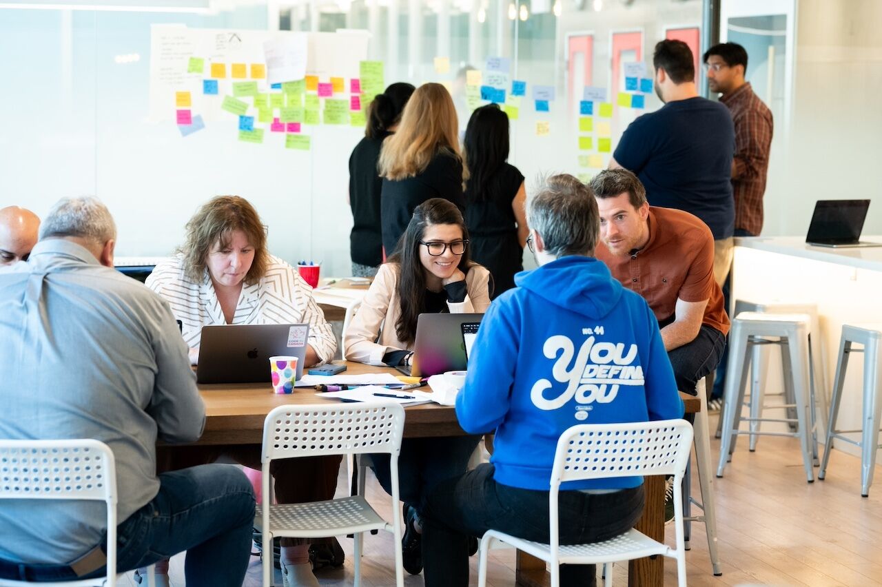 People working on laptops around a table with a group standing looking at wall of sticky notes in background